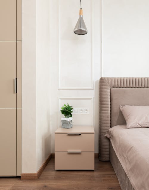 Bedside table with potted plant placed near comfortable bed with pink coverlet and pillows in cozy room at home