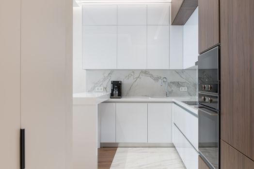Minimalist interior of small kitchen with white and wooden cupboards and modern appliances in light apartment