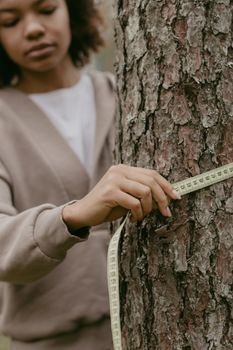 A focused young woman measures the circumference of a tree trunk using a tape measure outdoors.