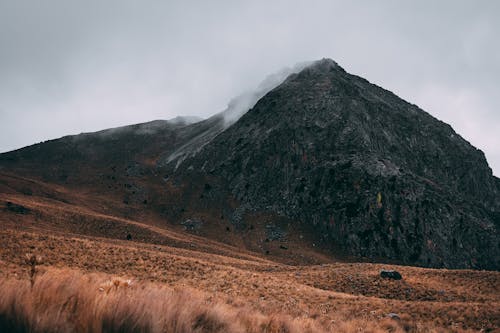 Brown Grass Field Near A Volcano Under Cloudy Sky