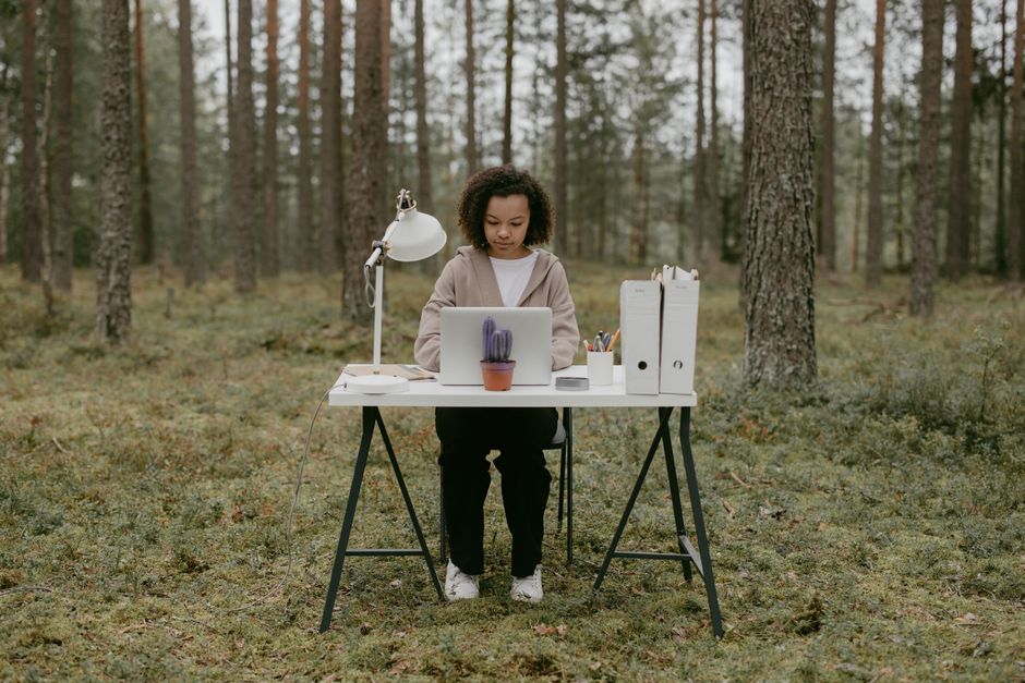 A woman sits at a desk in a forest, working remotely on a laptop amidst nature.