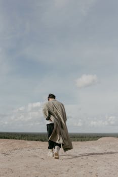 Back view of man walking in a vast desert landscape beneath a cloudy blue sky.