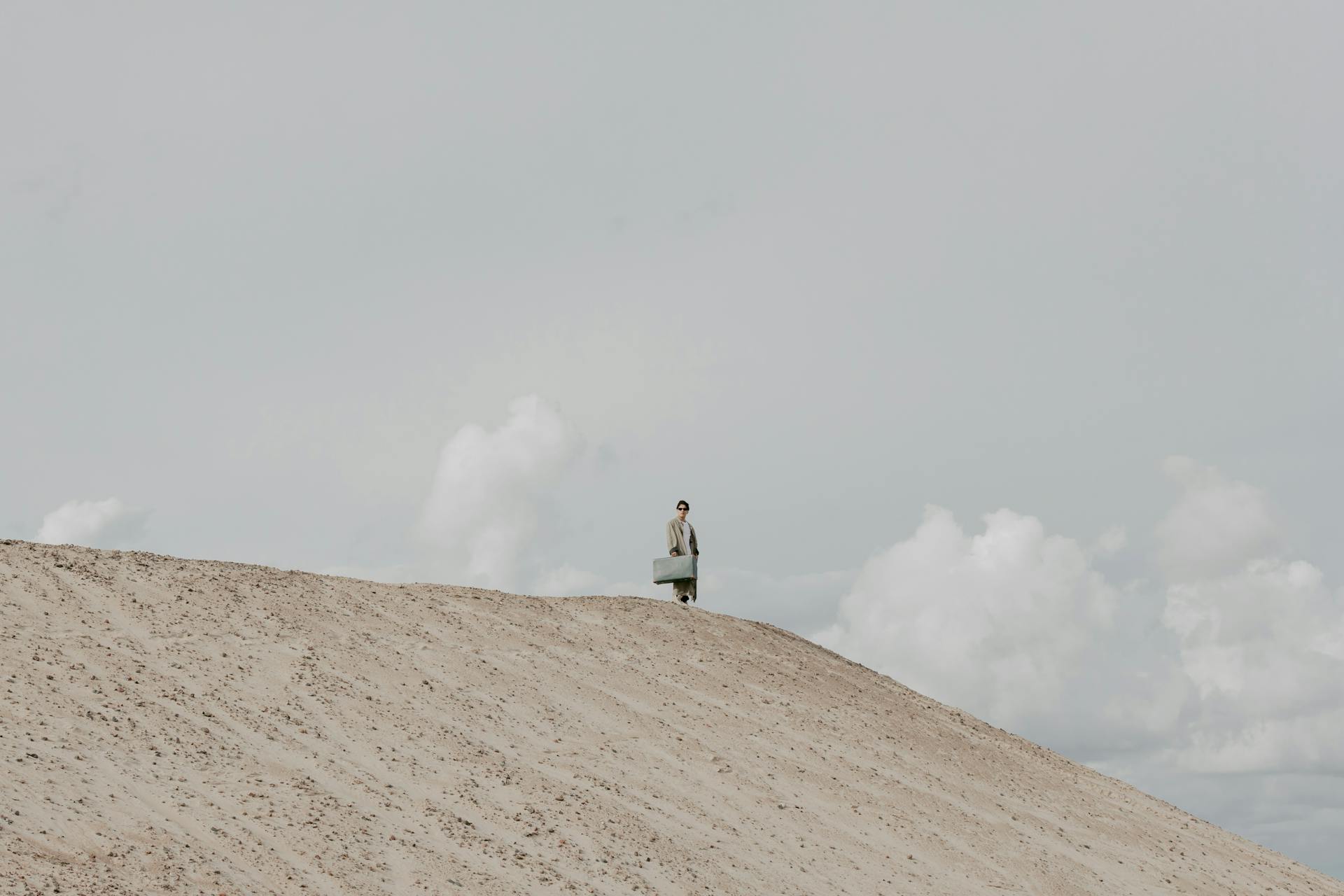 A lone traveler stands on a sandy hill with a suitcase under a cloudy sky.
