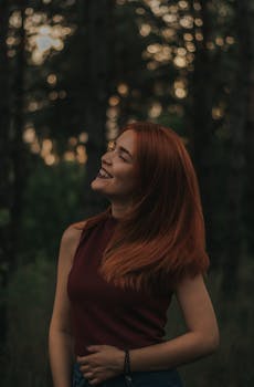 Smiling young woman in trendy outfit with makeup standing near trees with green leaves on grassy field in summer day in nature with closed eyes