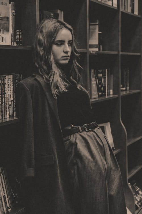 Black and white of thoughtful young female in trendy outfit leaning on bookshelf in library and looking away