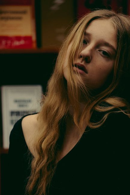Peaceful young female with long blond wavy hair leaning head on wall and looking at camera against bookshelf