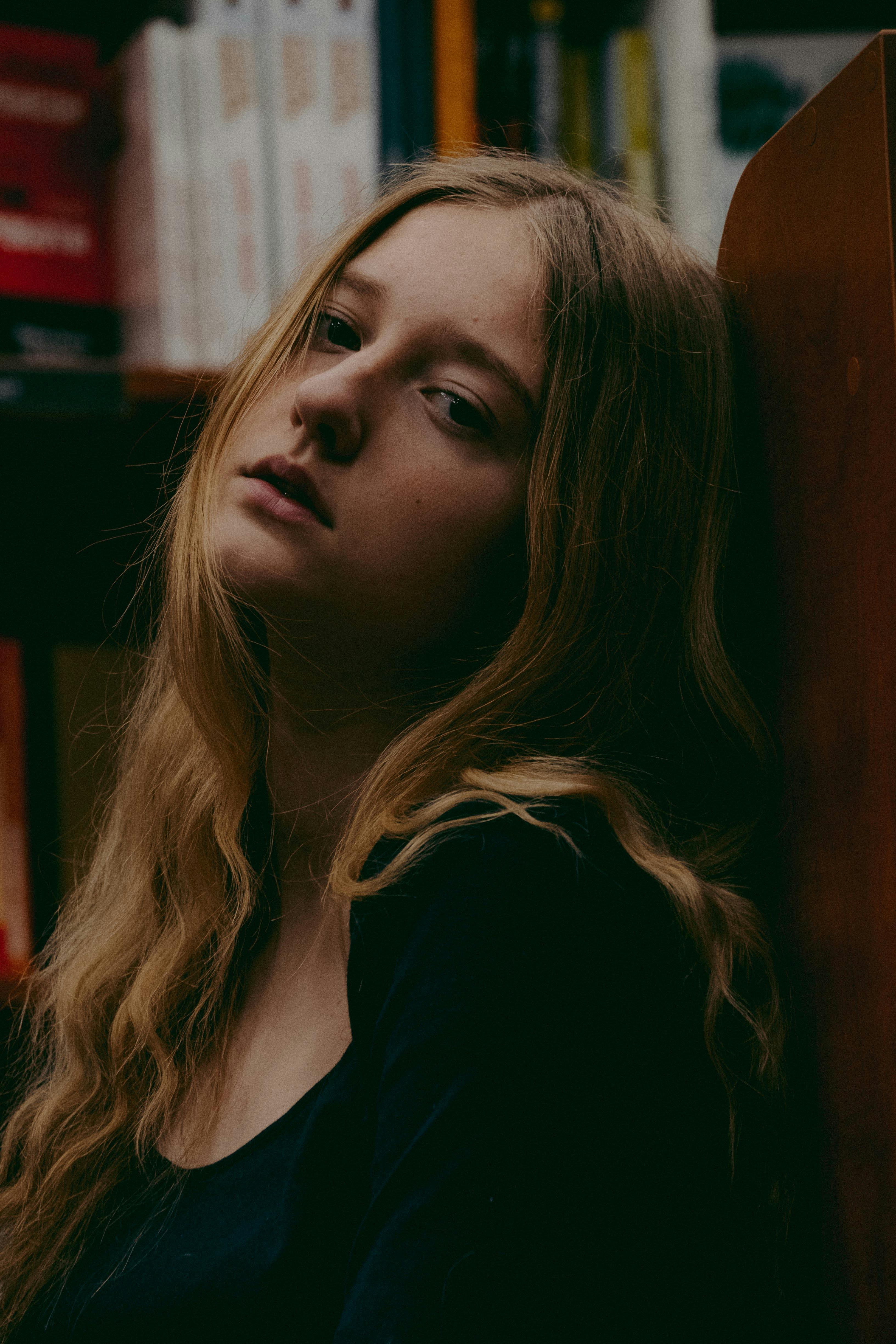 Side view of melancholic young female student with long blond wavy hair leaning on wooden bookshelf in library and looking at camera