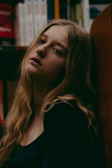 Side view of melancholic young female student with long blond wavy hair leaning on wooden bookshelf in library and looking at camera