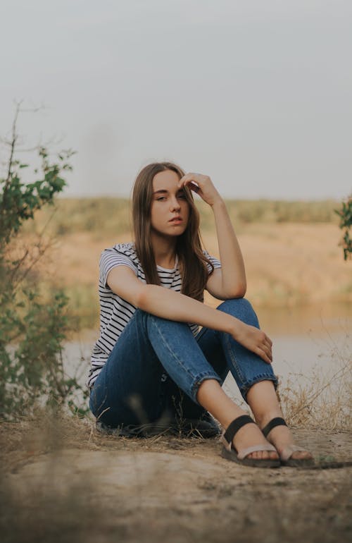 Full body of thoughtful young female millennial in casual outfit touching forehead while sitting on rural ground near meadow during holiday in countryside and looking at camera