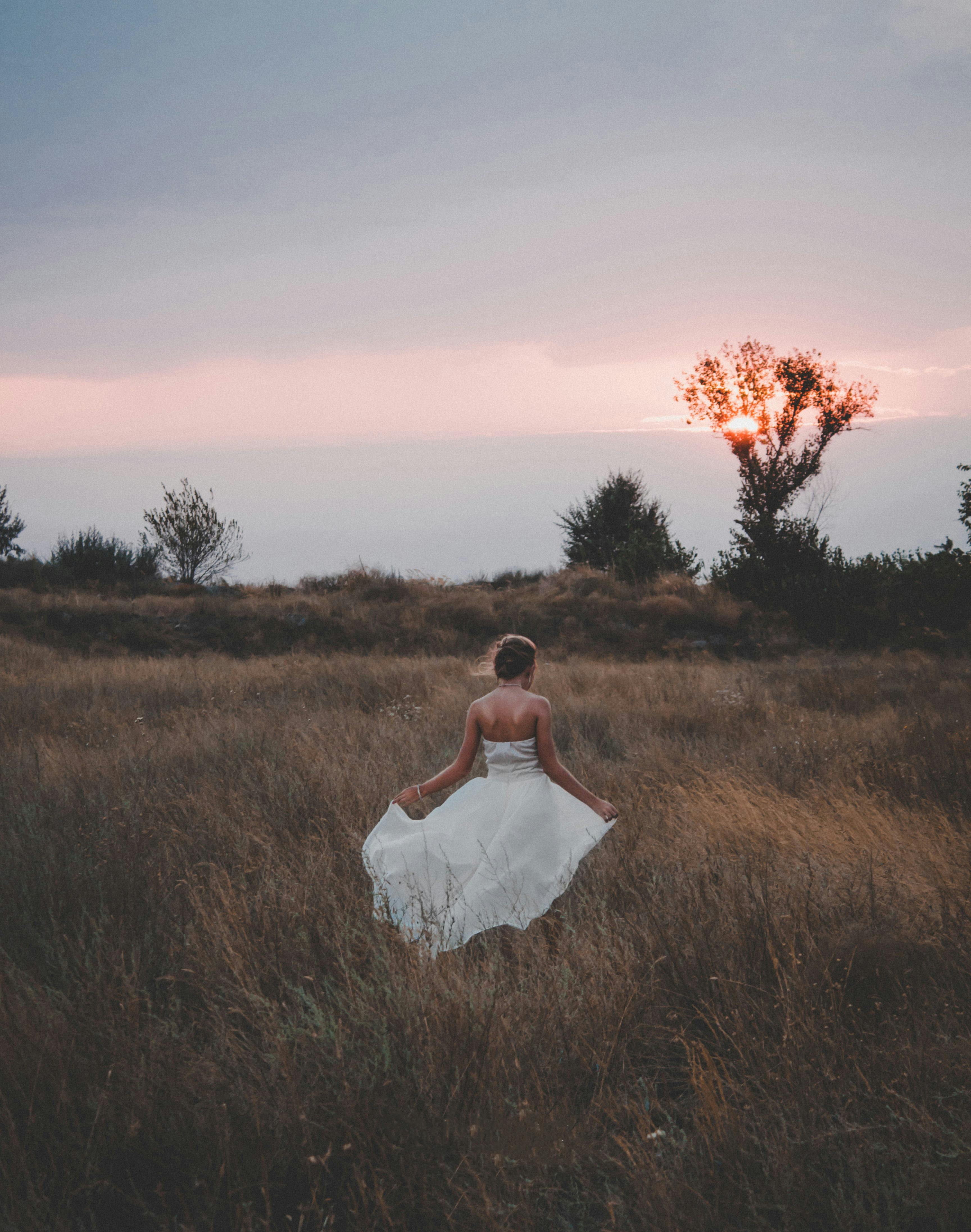Back view of unrecognizable bride in elegant white wedding dress walking on dry grassy meadow against cloudy sunset sky