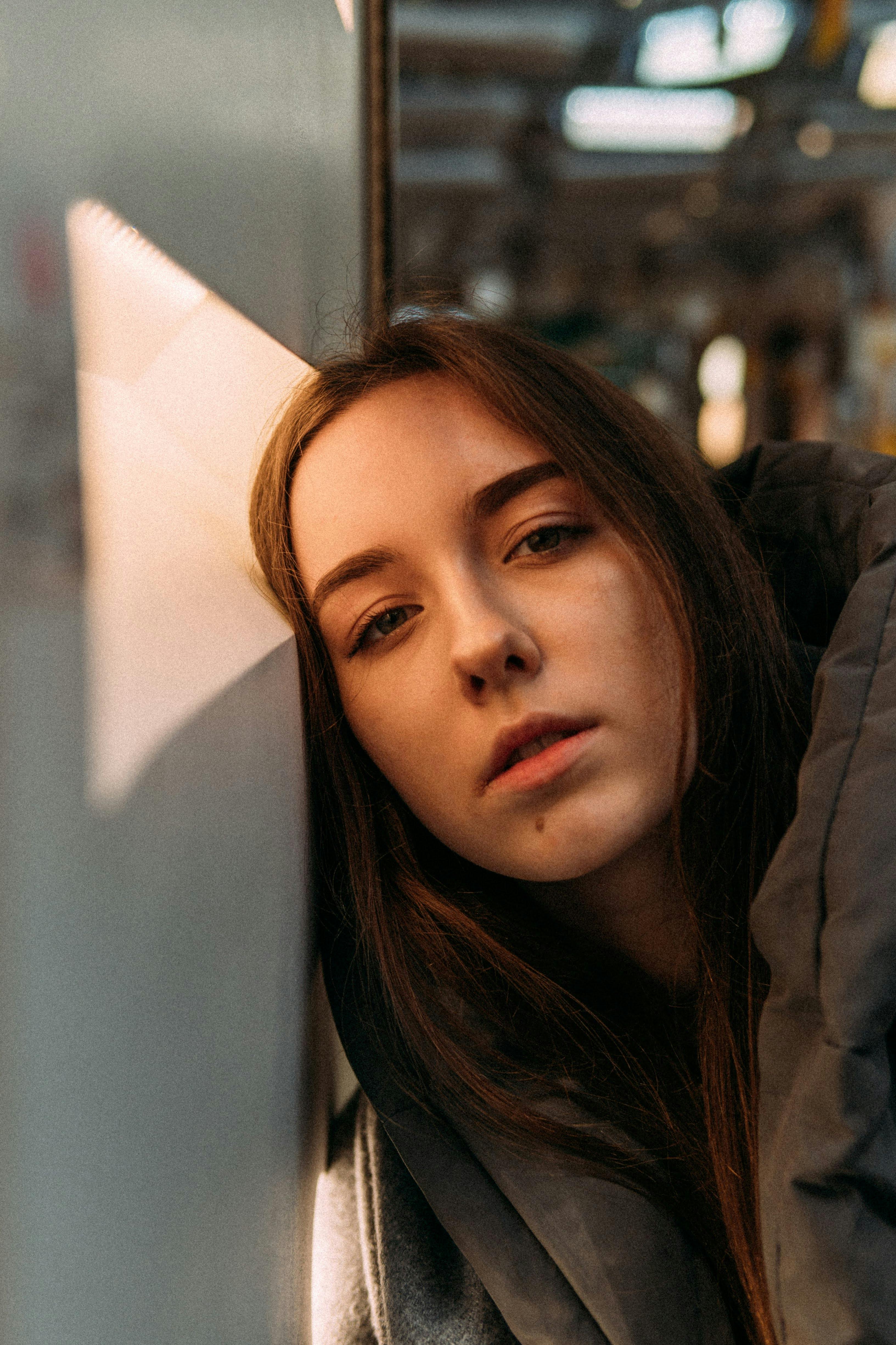 Pensive female teenager with dark hair in warm clothes leaning head on white wall and looking at camera in sunlight