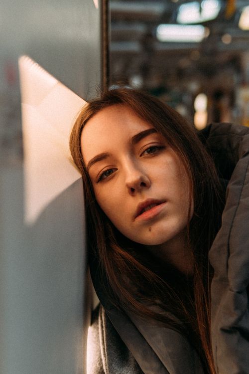 Free Pensive female teenager with dark hair in warm clothes leaning head on white wall and looking at camera in sunlight Stock Photo