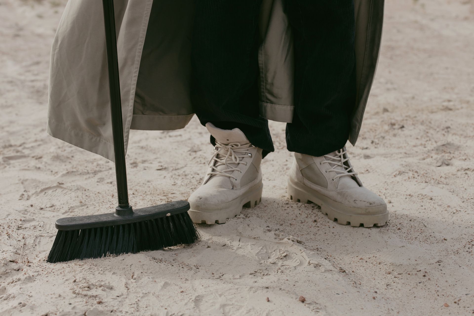 Close-up of a person wearing boots using a broom on sandy terrain, emphasizing footwear style and casual chores.