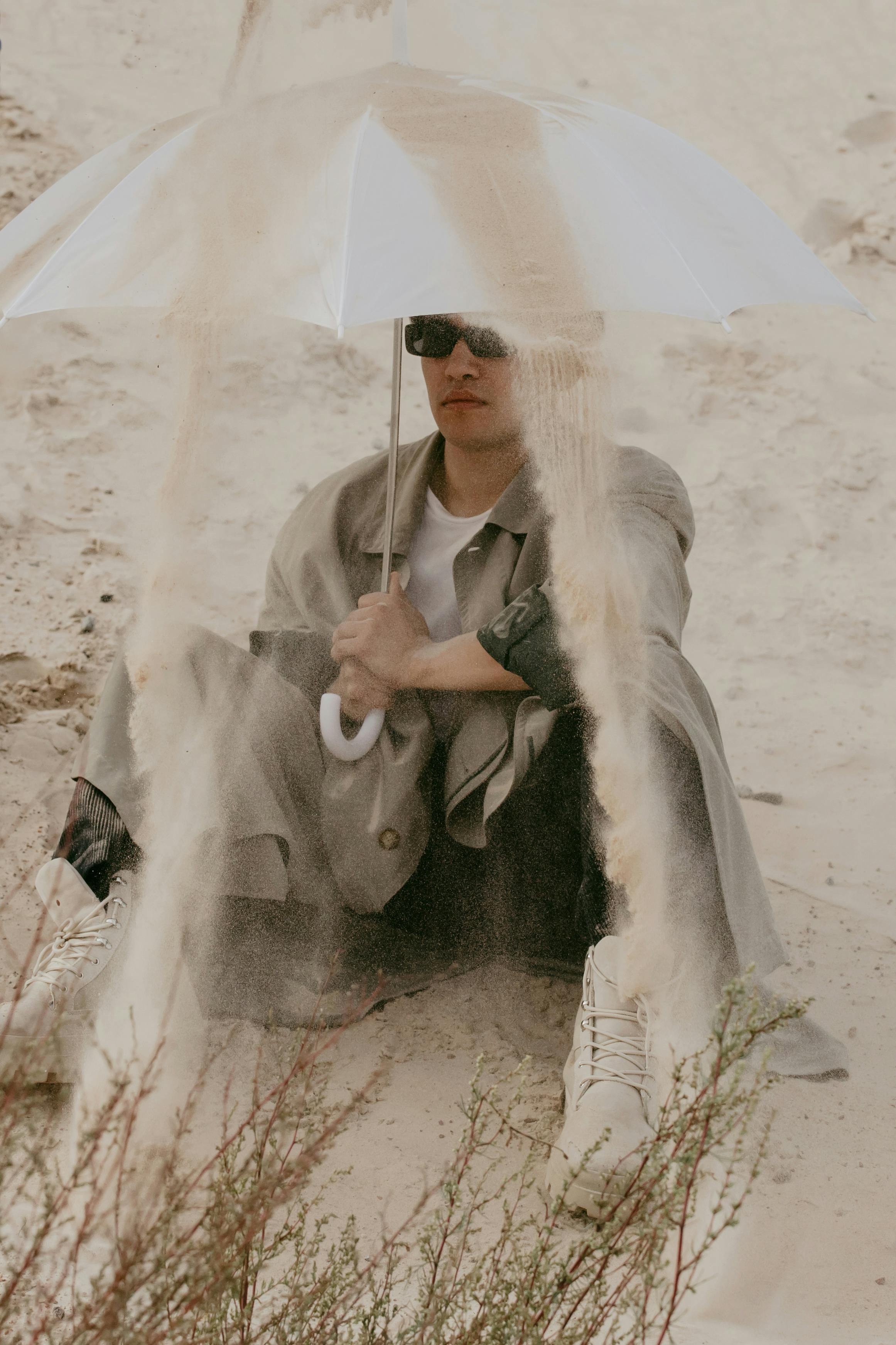 Stylish man in sunglasses sitting on sand holding an umbrella for shade in vibrant desert landscape.