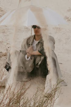 Stylish man in sunglasses sitting on sand holding an umbrella for shade in vibrant desert landscape.