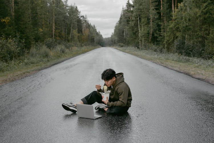 A Man In Brown Jacket Eating On The Road While Looking At His Laptop