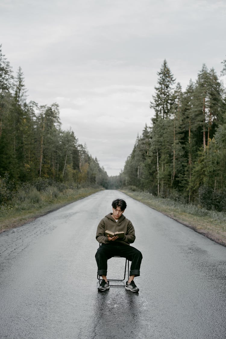 A Man In Brown Jacket Sitting On The Chair In The Middle Of The Road While Reading A Book