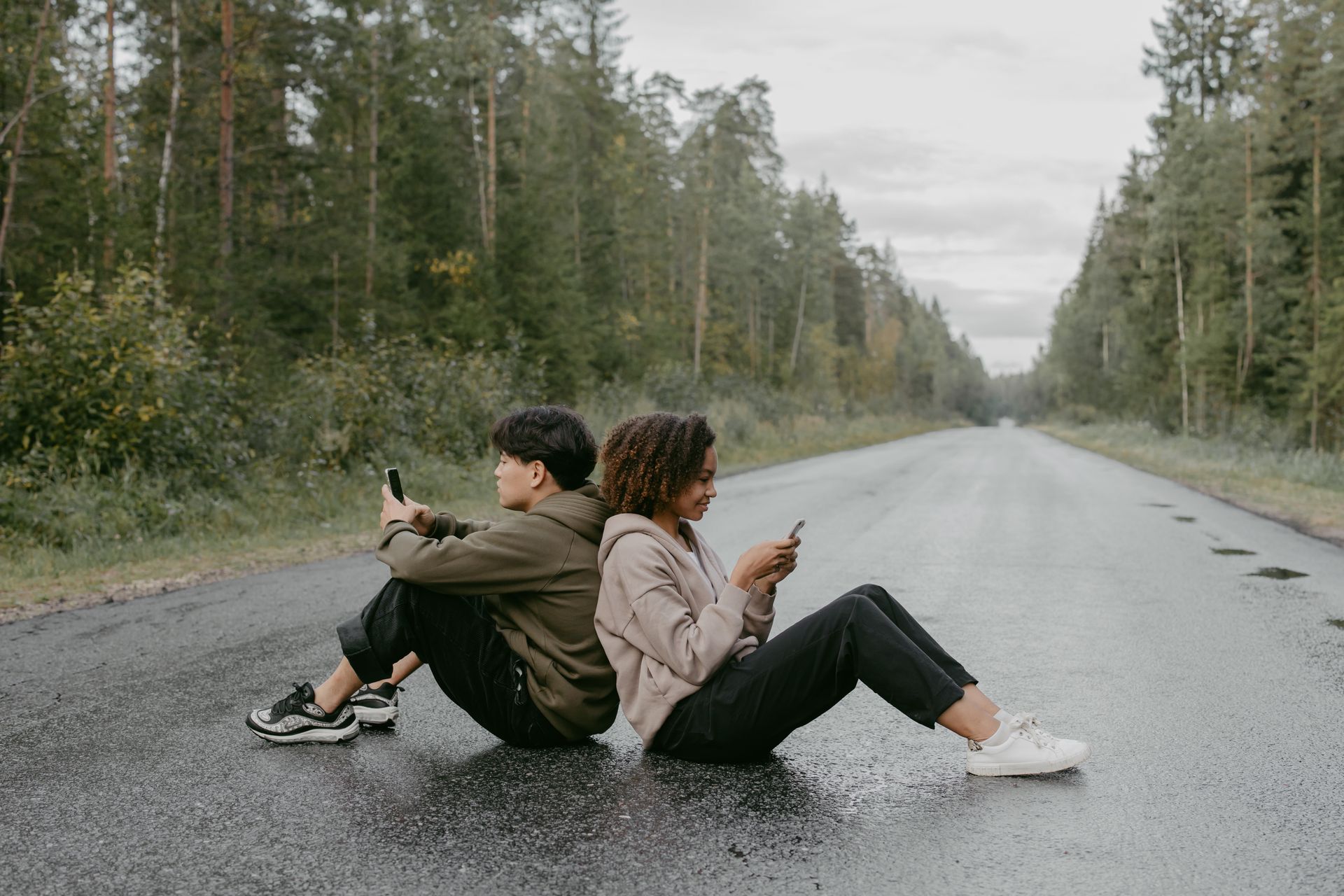 A young couple sitting back to back while texting on a remote forest road, showcasing modern communication.