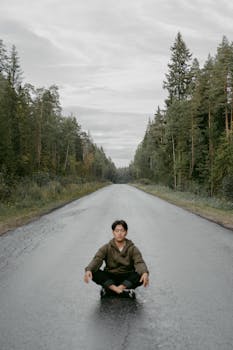 A man practicing yoga meditation on a quiet, wet road surrounded by forest.