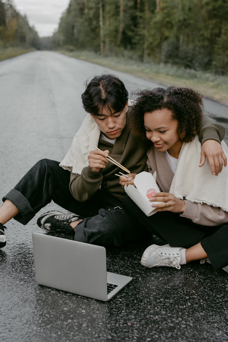 A Couple Sitting On A Wet Road Eating Food While Looking At The Screen Of A Laptop