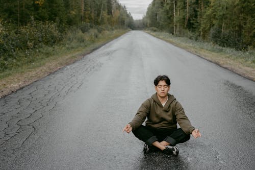 A Man Meditating on a Wet Road Between Green Trees