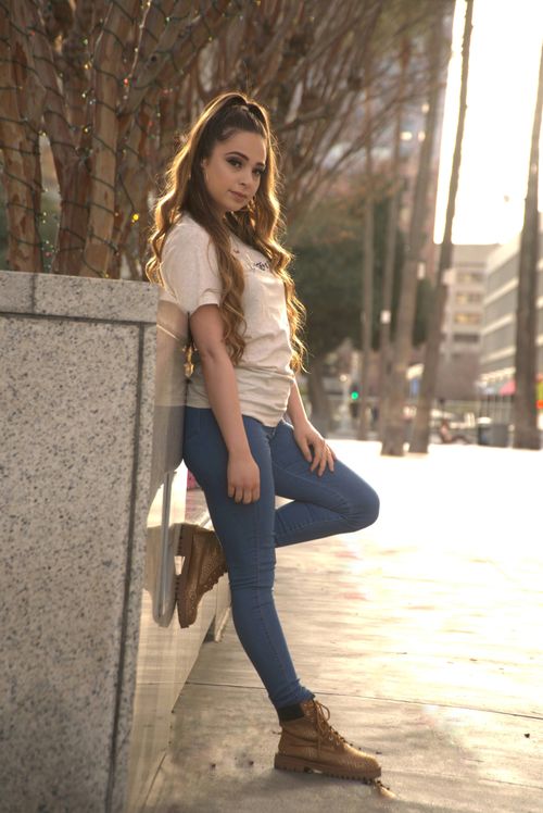 Free Full body side view of young female in t shirt and jeans leaning on stone fence near trees and buildings in city street and looking at camera in sunny day Stock Photo