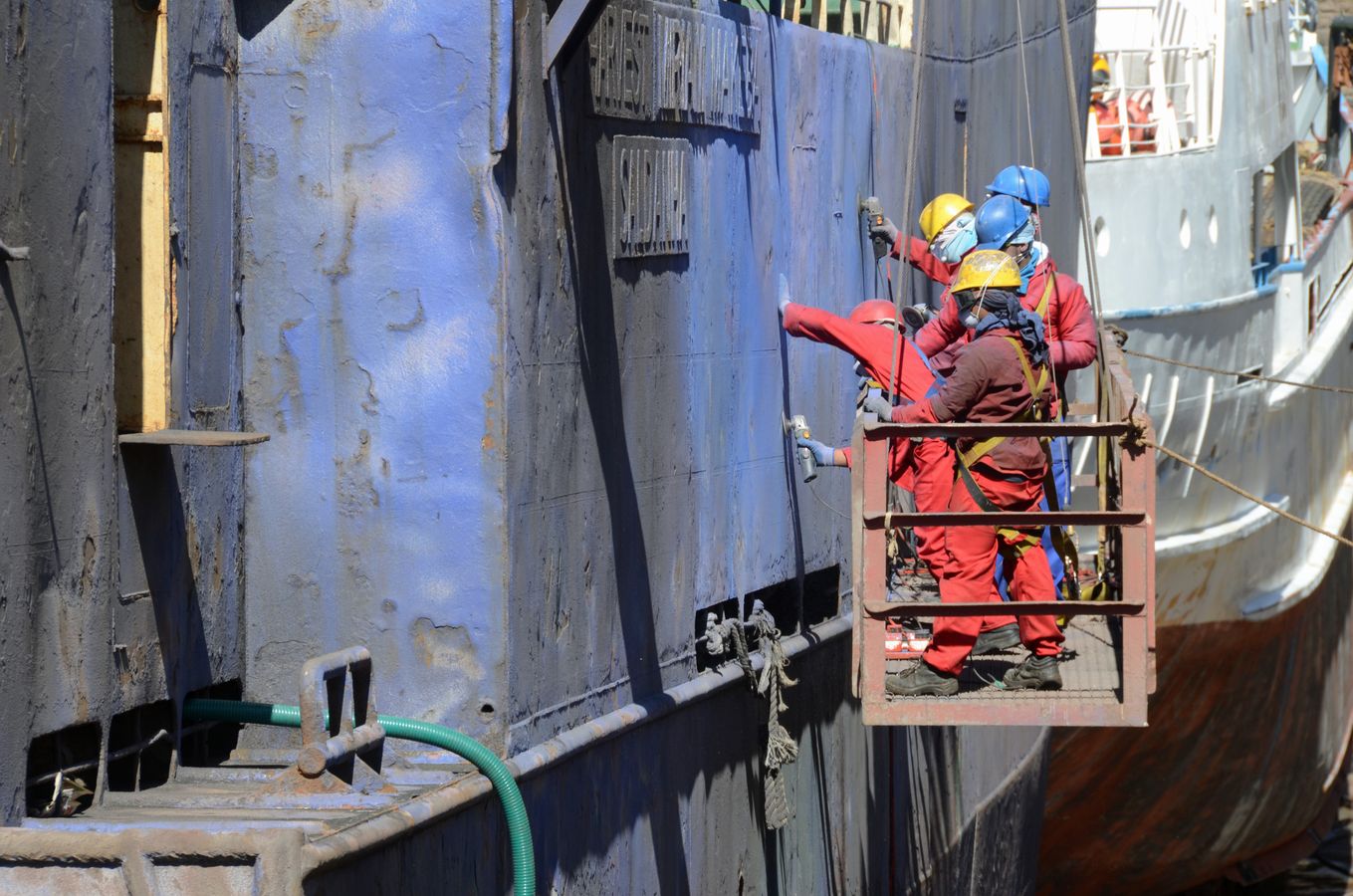 Workers on a suspended platform repairing the exterior of a ship in a shipyard.