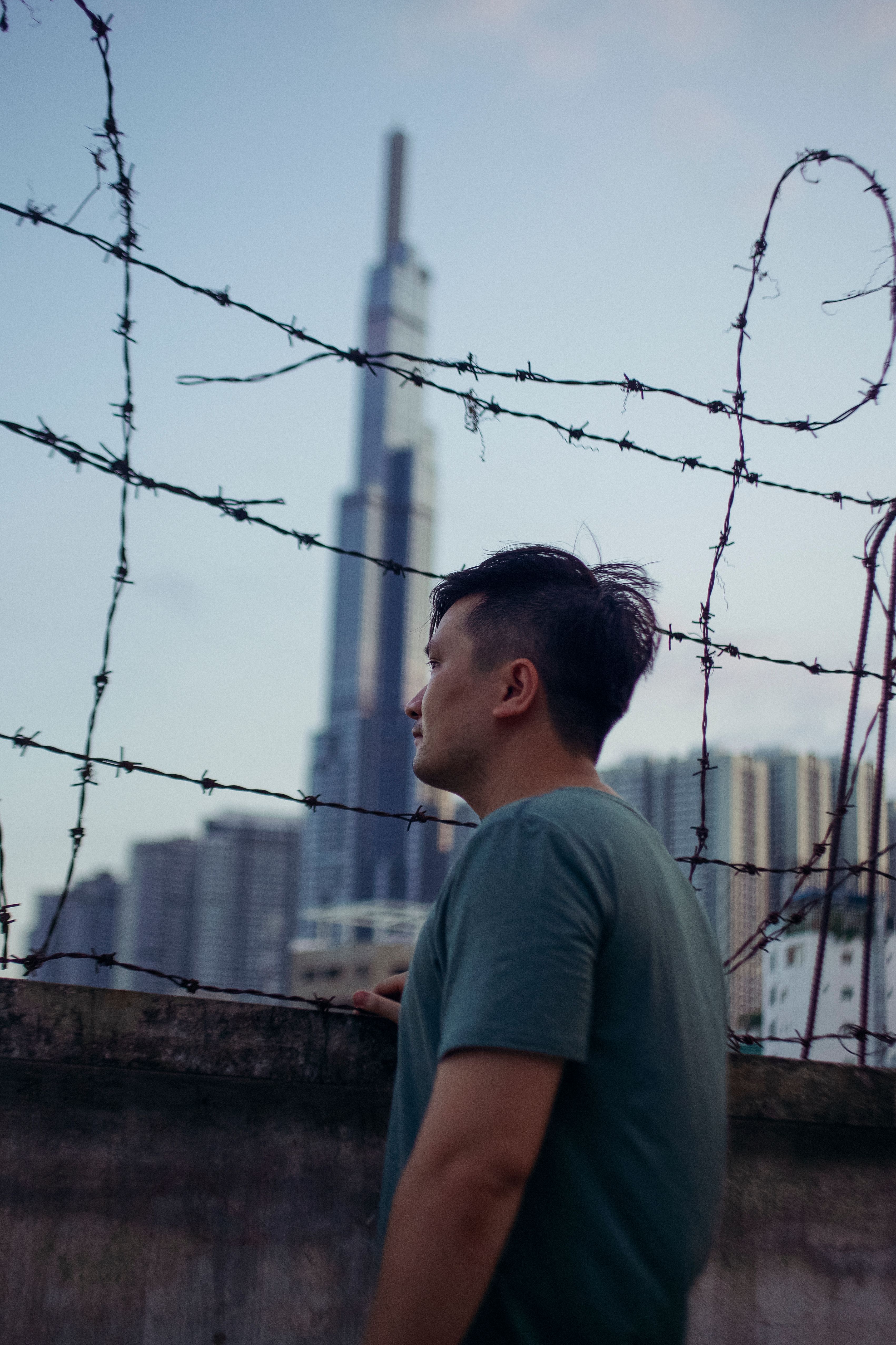 A man gazes through barbed wire towards a prominent skyscraper, depicting themes of isolation and urban life.