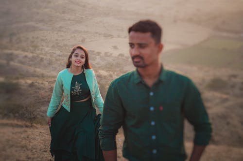 Positive Indian woman in long skirt looking at camera while standing on hilly terrain above grassy terrain with blurred boyfriend in nature