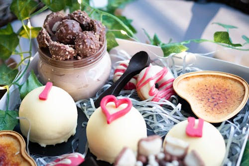 Tasty sweet little cakes covered with white chocolate and decorated with pink inscription placed in box near glass jar of chocolate sweets and doughnuts