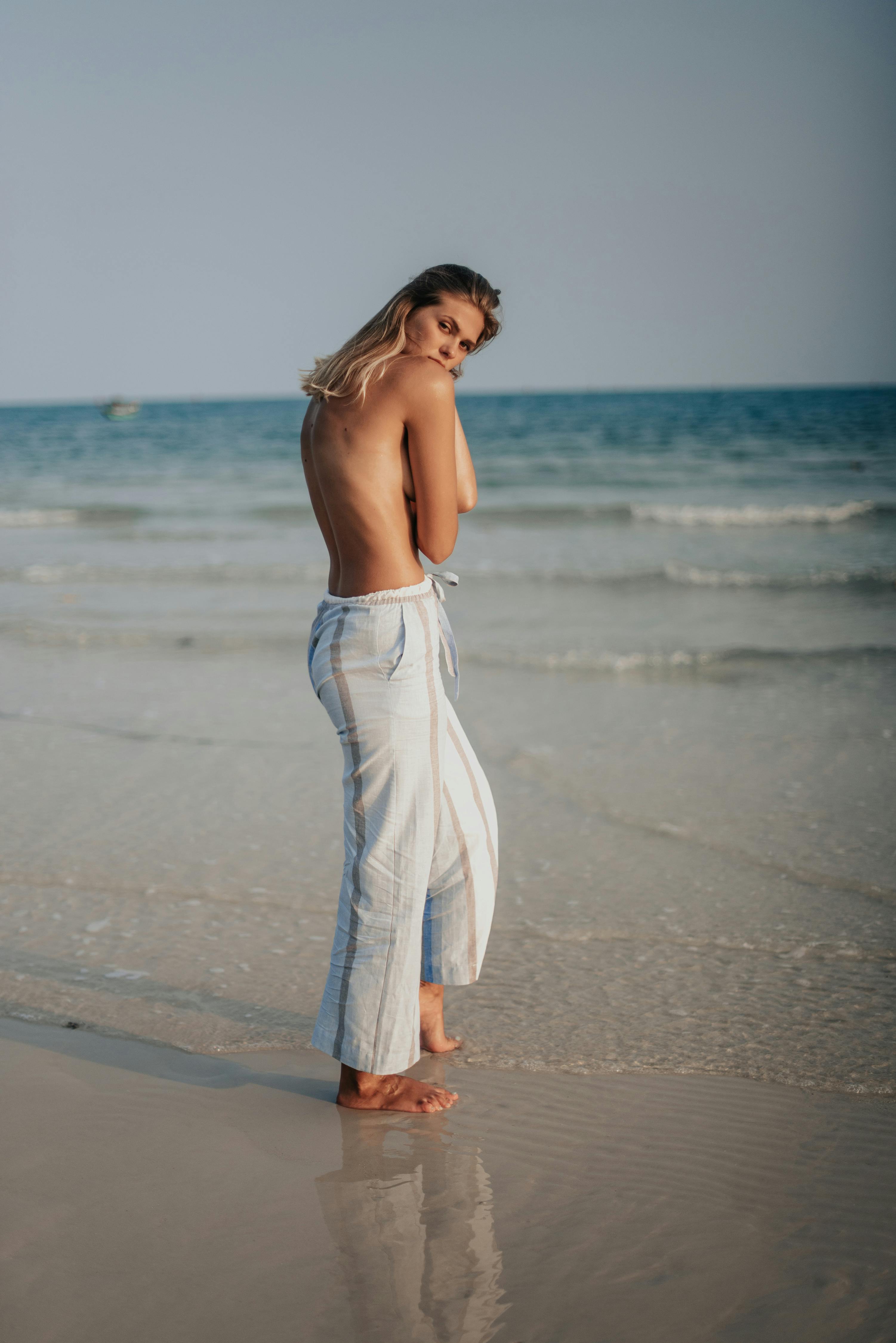 a topless woman in white pants standing on a beach