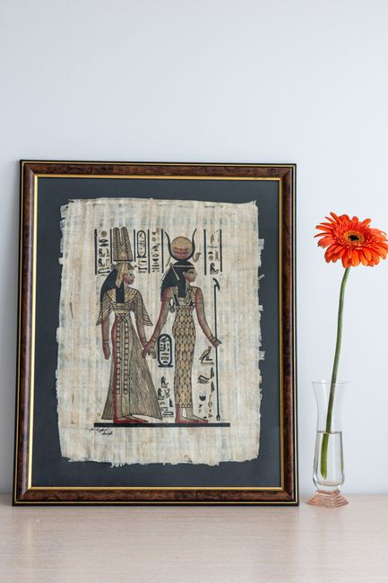 Ancient Egyptian artwork on papyrus alongside an orange gerbera in a glass vase.