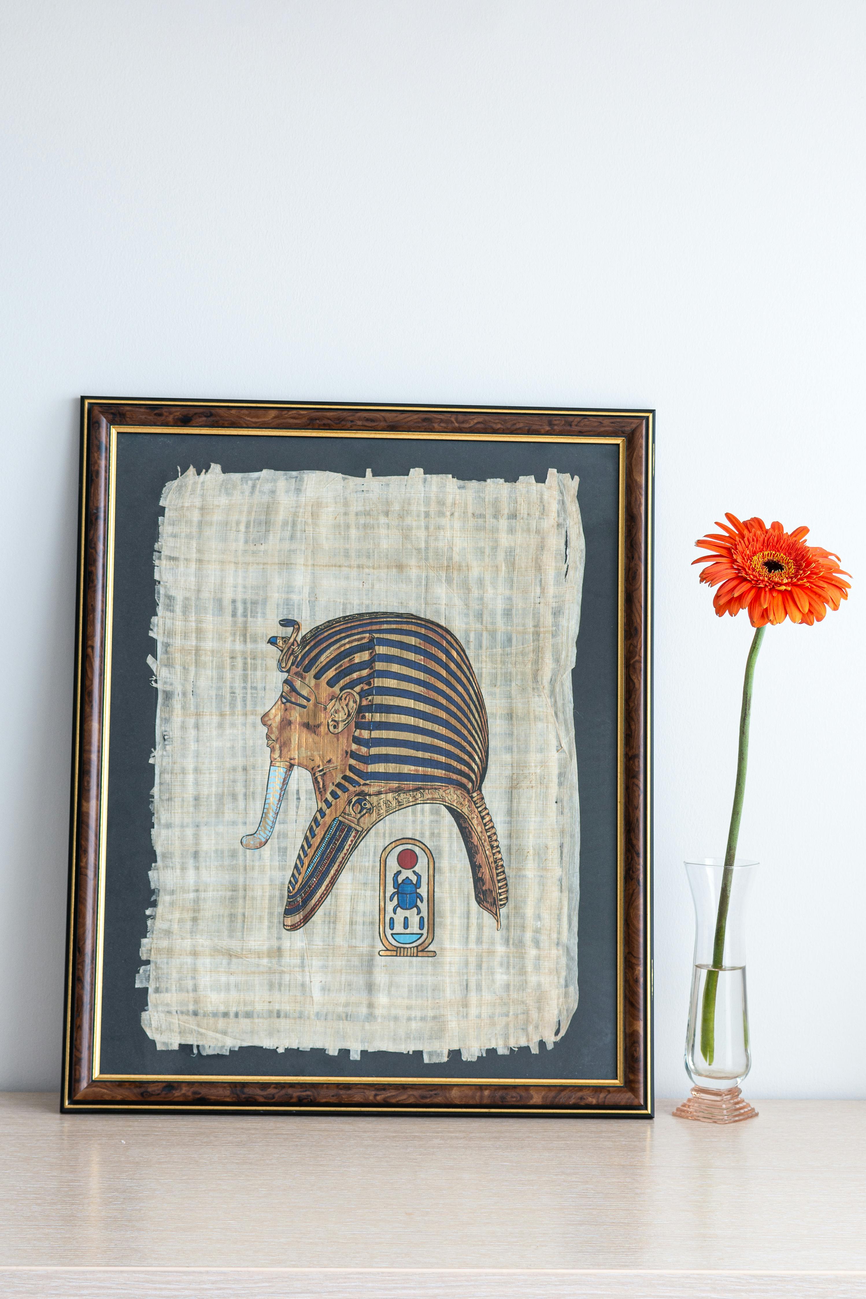 Classic framed papyrus painting of Tutankhamun with orange gerbera in a glass vase.