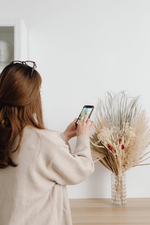 Woman in Beige Long Sleeve Shirt Taking Photo of Flowers in the Vase