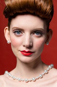 Close-up portrait of a woman with vintage hairstyle and pearl jewelry against a red background.