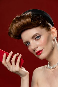 Elegant woman with a retro hairstyle holding a red phone, accentuated by pearl jewelry and a vibrant background.
