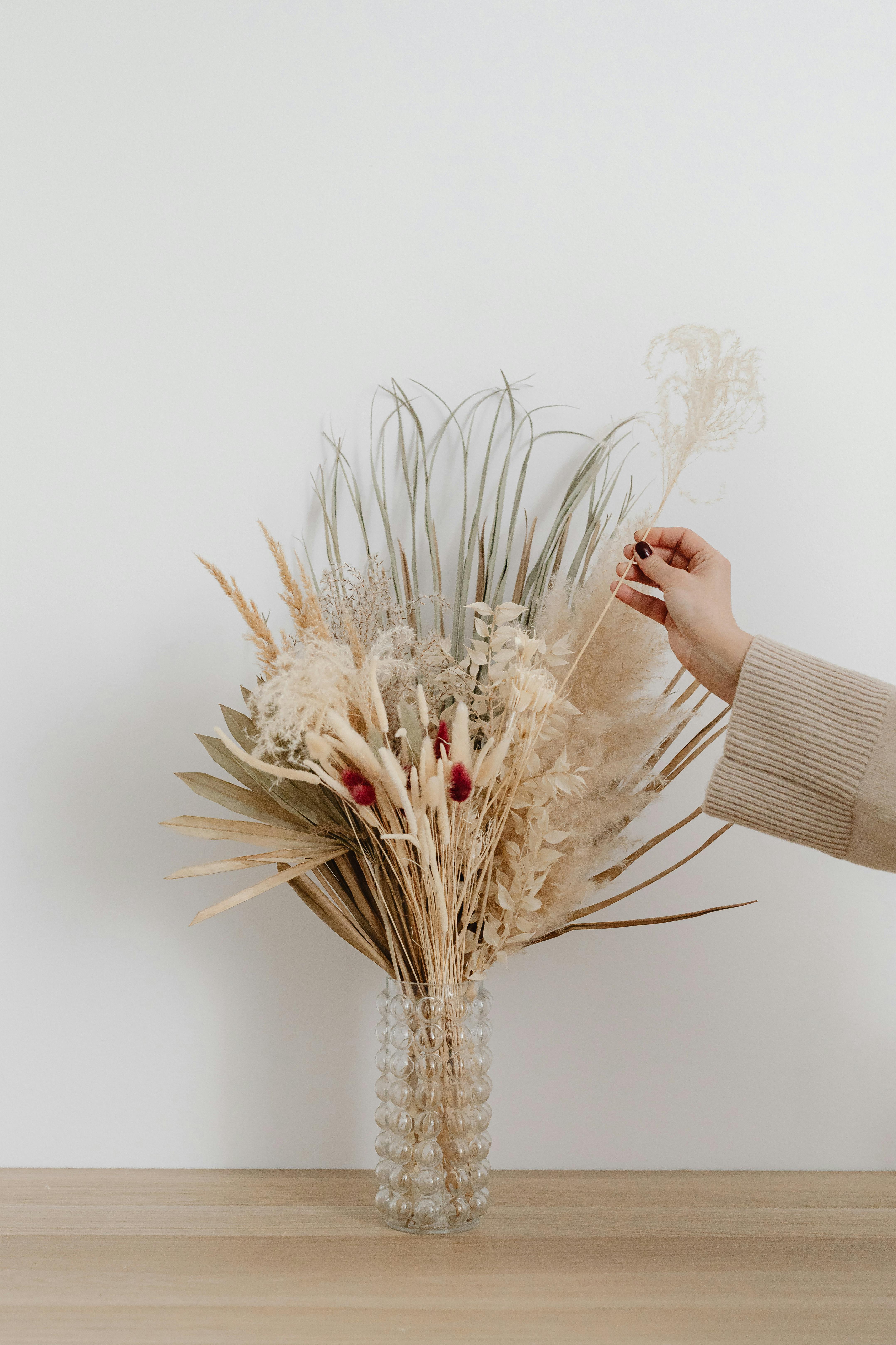 Artistic arrangement of dried flowers in a glass vase with a hand adjusting the bouquet on a white background.