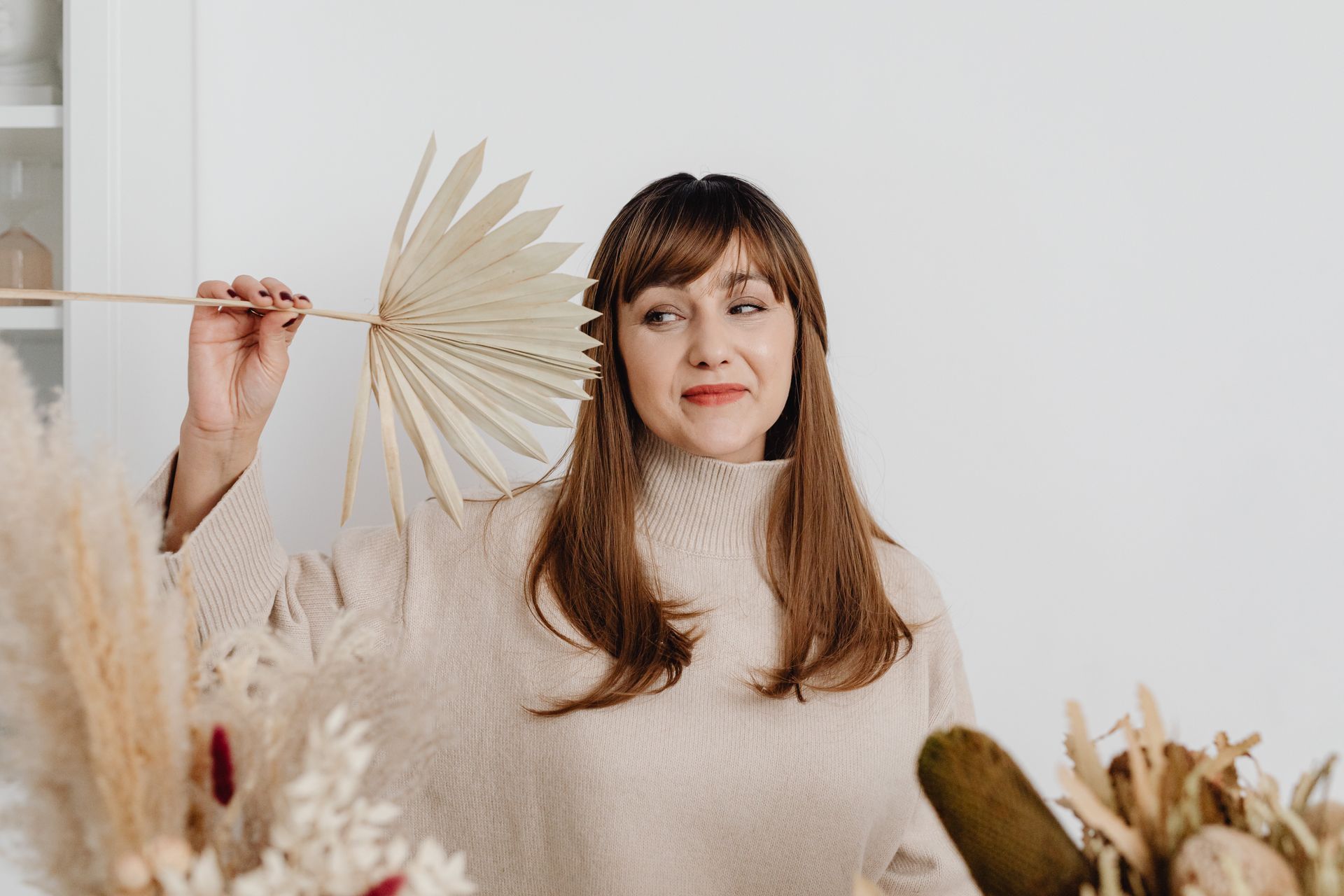 A smiling woman in a beige sweater holding a paper fan surrounded by autumn decor indoors.