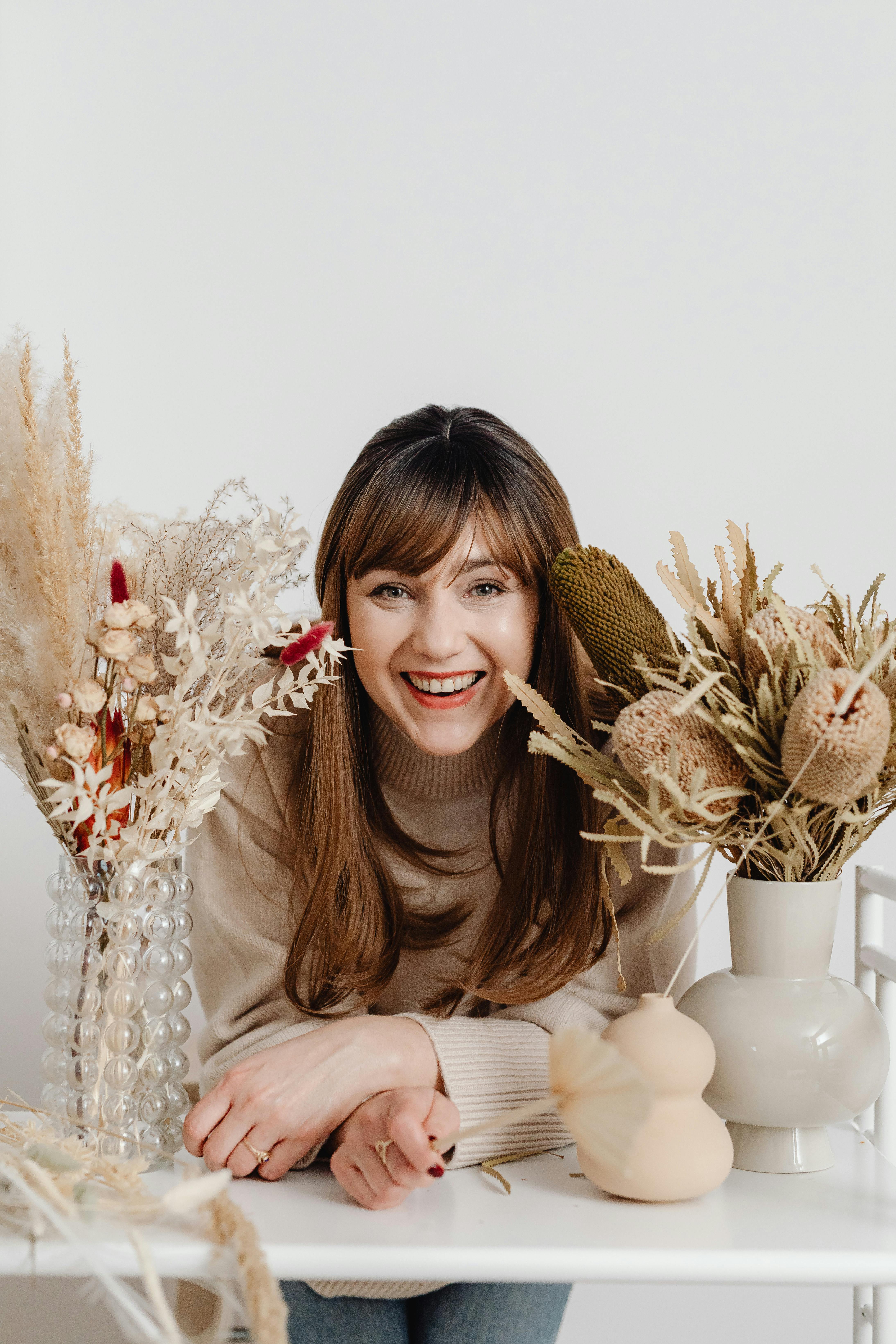 Smiling woman with long hair surrounded by dried flowers and vases indoors, exuding warmth and creativity.