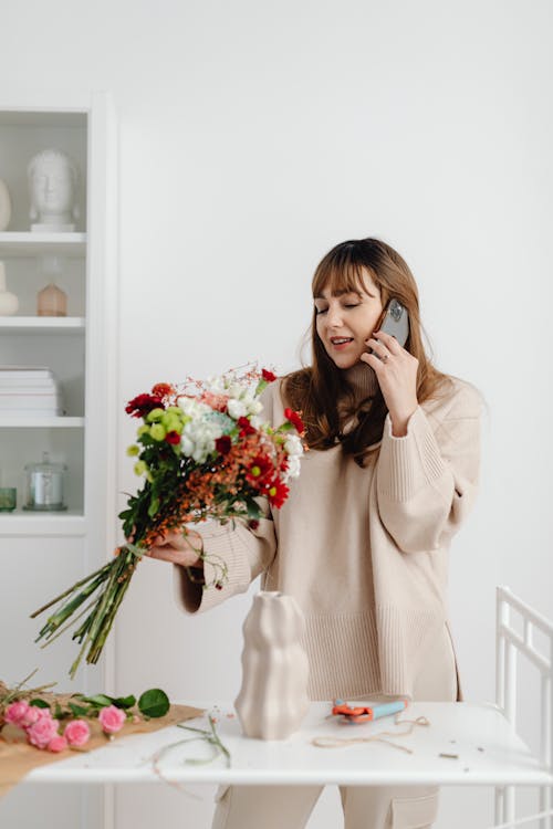 Woman in Beige Long Sleeve Shirt Holding a Bouquet of Flowers while Talking on the Phone