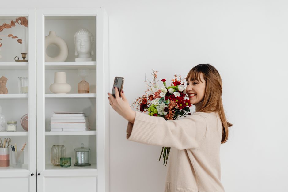 Smiling woman holding a bouquet and taking a selfie in a cozy indoor setting.