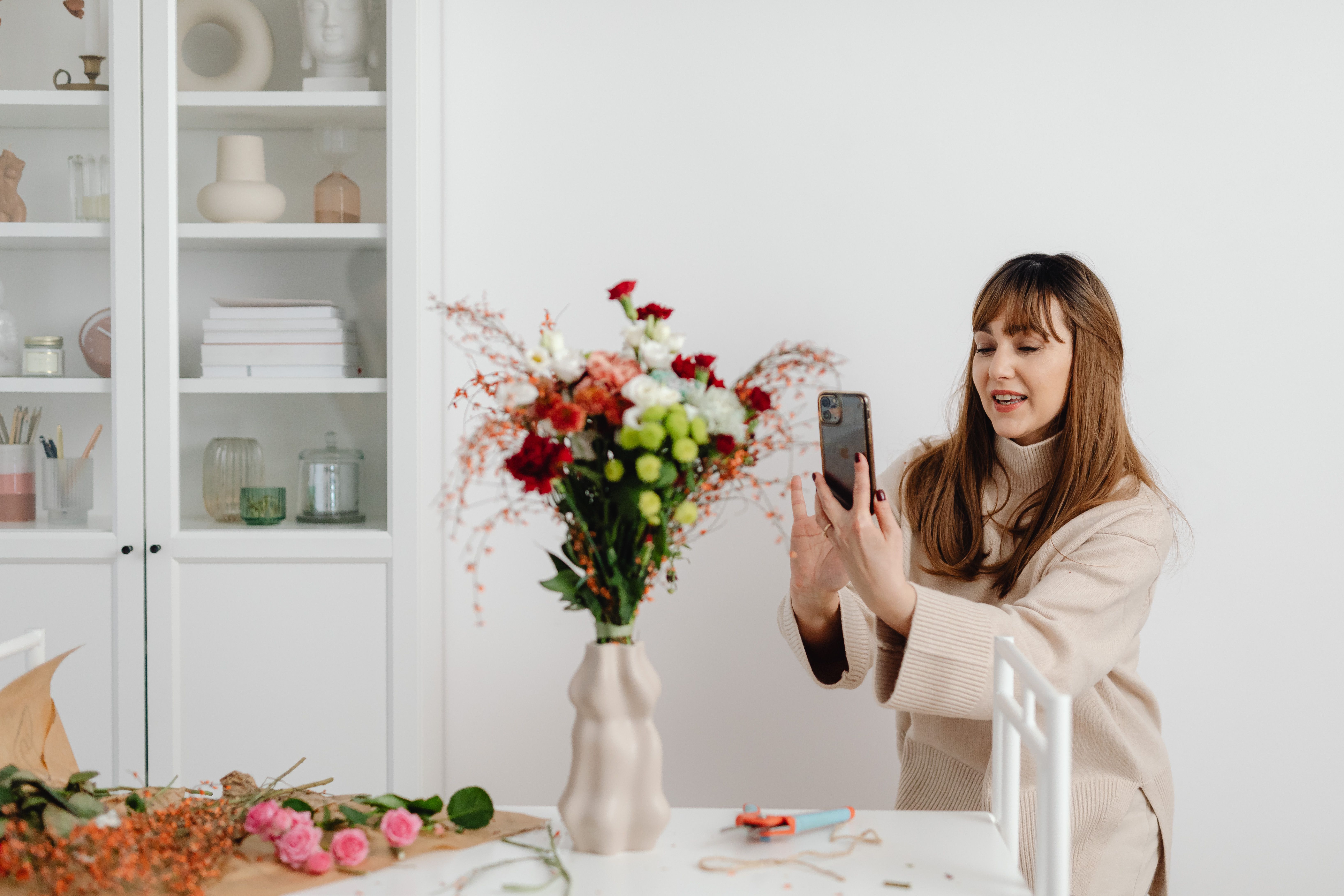 A woman takes a photo of her artistic flower arrangement indoors with a smartphone.