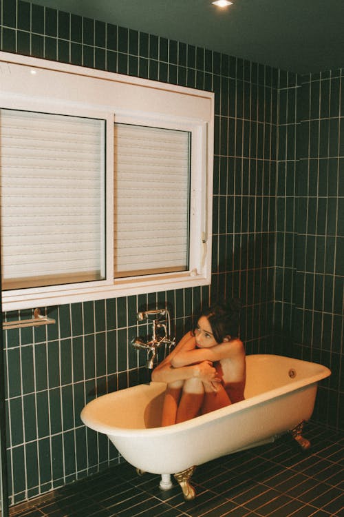 High angle of frightened nude female hiding bare body while sitting in bathtub and looking away