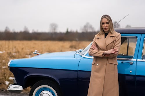 Pensive female in stylish clothes standing with crossed arms near aged vintage car and looking at camera in countryside in daytime