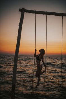 A woman in swimwear poses on a swing over the ocean at sunset, creating a stunning silhouette.