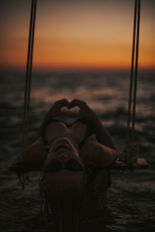 Free A woman in sunglasses reclines on a swing above the ocean at sunset, creating a serene silhouette. Stock Photo