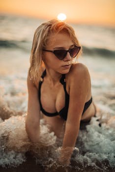 A young woman posing in the surf at sunset on a beach, wearing sunglasses.