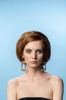 Close-up portrait of a stylish woman with red hair and oversized earrings on a blue background.