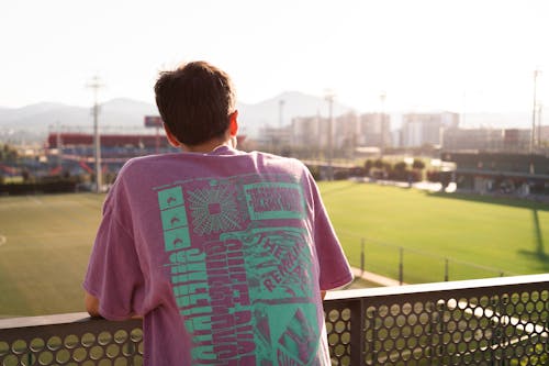 Back view of anonymous young male in colorful t shirt standing leaning on metal fence on stadium and looking at play filed