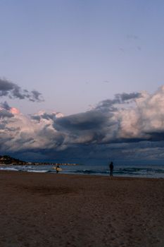 Picturesque scenery of sandy beach with surfers near foamy sea under cloudy sky in evening